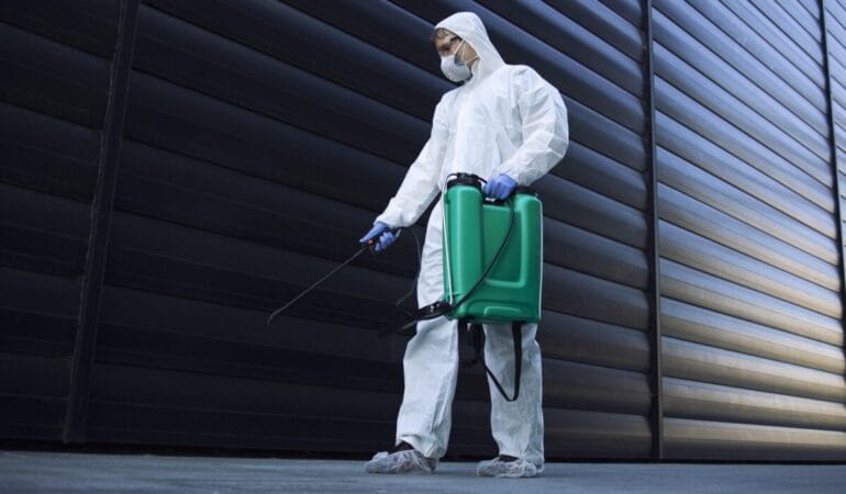 Person in white chemical protection suit doing disinfection of public areas to stop spreading highly contagious corona virus.
